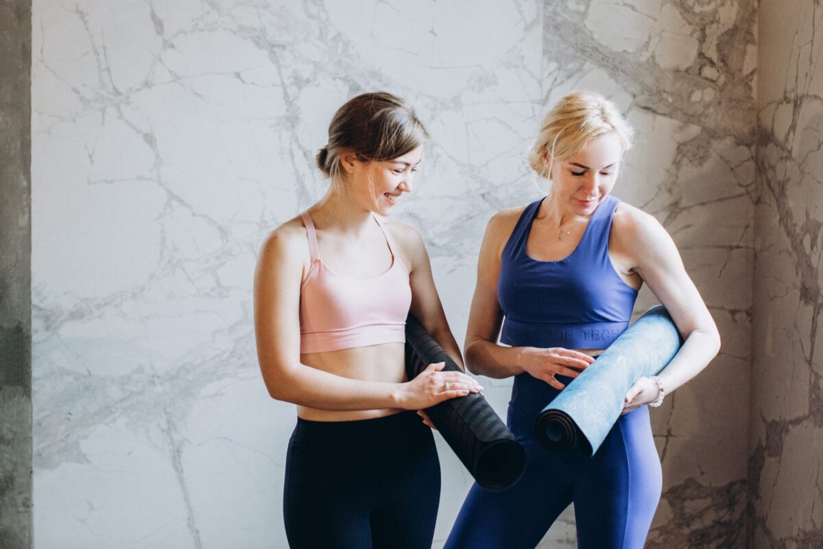 Two women in activewear holding yoga mats, smiling and preparing for a fitness session.